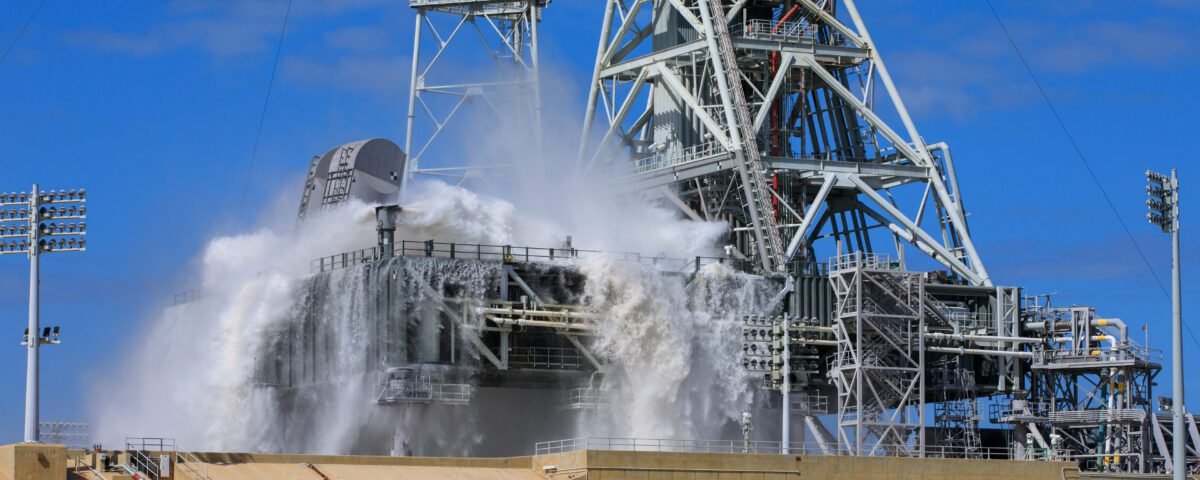 A large amount of water cascades over the edges of the gray mobile launcher at NASA's Kennedy Space Center. Droplets of water also spray up through the air, creating a mist.
