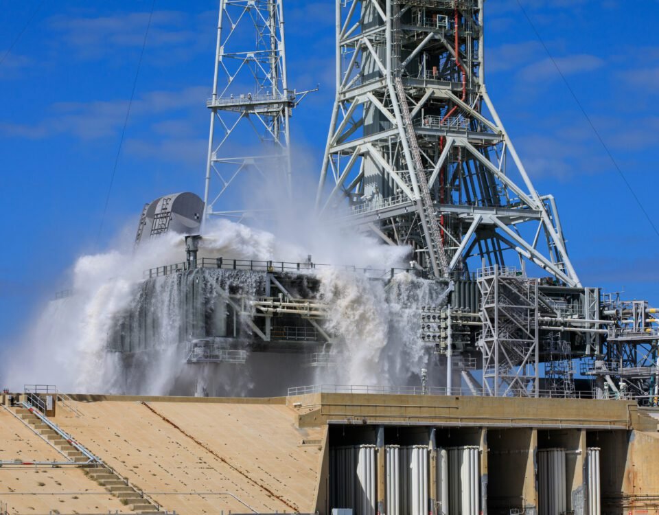 A large amount of water cascades over the edges of the gray mobile launcher at NASA's Kennedy Space Center. Droplets of water also spray up through the air, creating a mist.