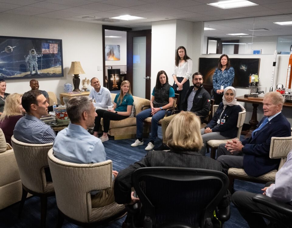 A group of 17 people sit and stand in a circle in a meeting area in an office. There are rocket models and space-themed imagery decorating the space. Everyone looks toward NASA administrator Bill Nelson, second from right, as he speaks.