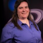 Lynn Bassford, a woman with long brown hair, smiles at the camera in an official headshot. She wears a purple collared shirt and poses in front of a photo of Saturn and Neptune.