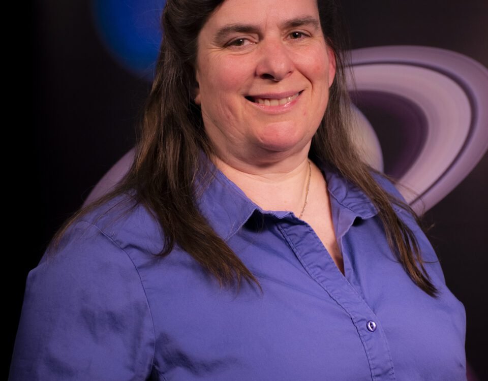 Lynn Bassford, a woman with long brown hair, smiles at the camera in an official headshot. She wears a purple collared shirt and poses in front of a photo of Saturn and Neptune.