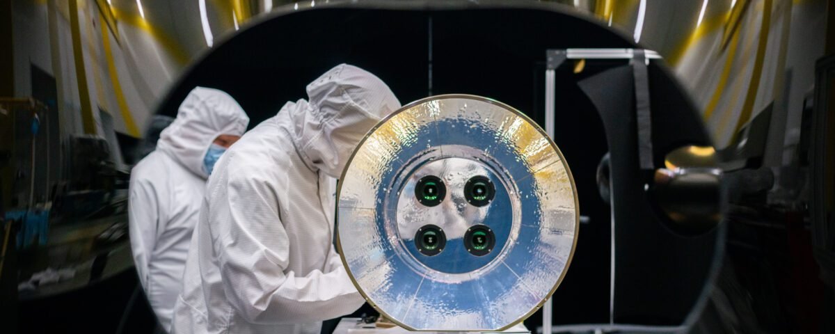 Two scientists wearing white lab coats, hoods, and masks work behind a large reflective disc with four camera lenses in its center, part of the assembly for the AWE.