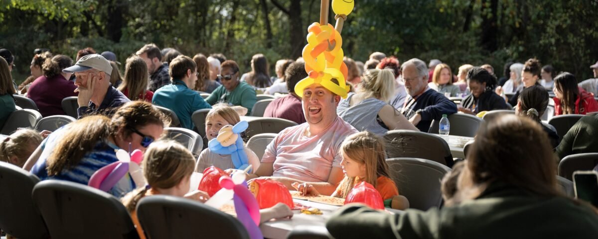 A man with an open-mouthed smile wears a balloon hat made at the Fall Family Fest alongside children who are also enjoying their balloon toys during a Bingo round.