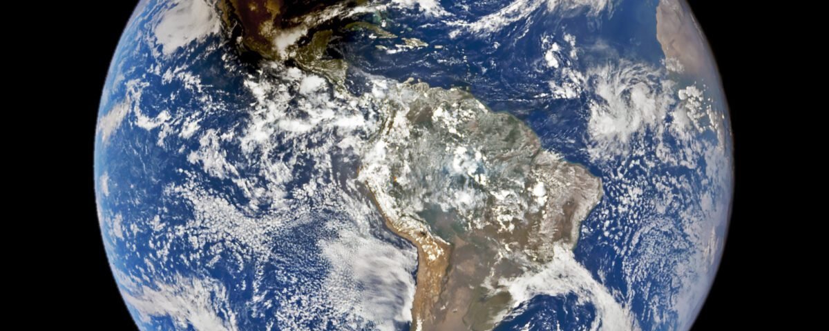 A full disc image of Earth in which North and South America are visible. The Moon casts a dark brown shadow in the top left quadrant.