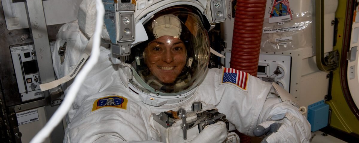 Astronaut Nicole Mann faces the camera and smiles. She is wearing a white spacesuit, which has an American flag patch on the left arm, and another patch on the right arm.