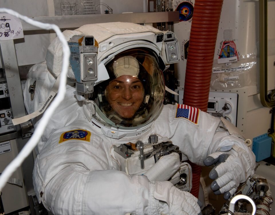 Astronaut Nicole Mann faces the camera and smiles. She is wearing a white spacesuit, which has an American flag patch on the left arm, and another patch on the right arm.