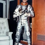 Astronaut Alan Shepard smiles while wearing an all-silver pressure suit for the Mercury mission. He holds a white helmet in his hands.