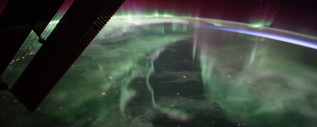 A green, fog-like haze stretches over Canada. At the edges of Earth’s atmosphere, the light takes on a purple-red color, eventually fading into the darkness of space. Some stars are faintly visible. At top left, a portion of the solar arrays of the International Space Station is backlit against the planet.