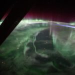A green, fog-like haze stretches over Canada. At the edges of Earth’s atmosphere, the light takes on a purple-red color, eventually fading into the darkness of space. Some stars are faintly visible. At top left, a portion of the solar arrays of the International Space Station is backlit against the planet.