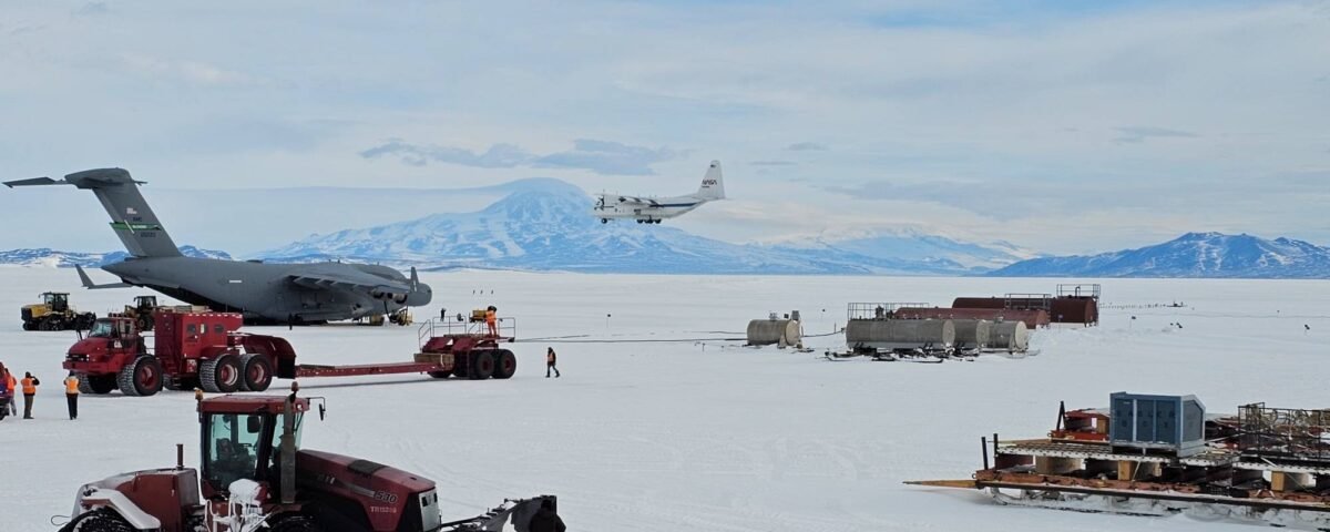 Tractors and other heavy machinery rest on the snowy plain in the foreground. In the middle of the image, a dark gray aircraft sits on the ground as a smaller white aircraft, NASA's C-130, lands.