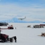 Tractors and other heavy machinery rest on the snowy plain in the foreground. In the middle of the image, a dark gray aircraft sits on the ground as a smaller white aircraft, NASA's C-130, lands.