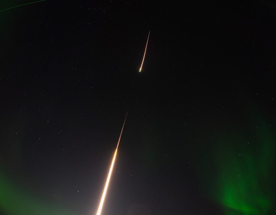 A long-exposure photograph of a sounding rocket launching in a night sky highlighted by aurora. The sounding rocket is a bright white streak, leaving from a snow-covered ground and moving up into the sky. A small break in the streak represents the first stage of the rocket burning out and the second stage igniting. A soft, green aurora frames the edges of the image, with several white stars speckled through the black sky. A bright green line toward the top of the frame represents a LIDAR beam. A fisheye lens was used for the photograph, creating a curve for the ground and LIDAR beam.