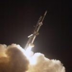 a 3 stage sounding rocket launches off a rail against a inky black sky. In the foreground plumes of white smoke are lit up by the rocket's ignition