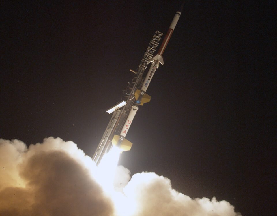 a 3 stage sounding rocket launches off a rail against a inky black sky. In the foreground plumes of white smoke are lit up by the rocket's ignition