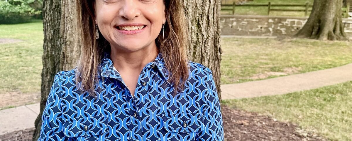 Rita Owens, a woman with shoulder-length, light brown hair, smiles at the camera in a casual outdoor portrait. She wears a patterned blue dress and stands in front of a tree. More green trees, a stone wall and brick buildings with cars are visible in the background.