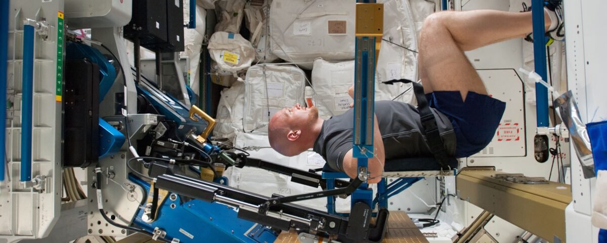 ESA astronaut Alexander Gerst in a squat position while working out on the ARED, with his arms against a beam. His body is facing to the right and his head turned to smile at the camera.