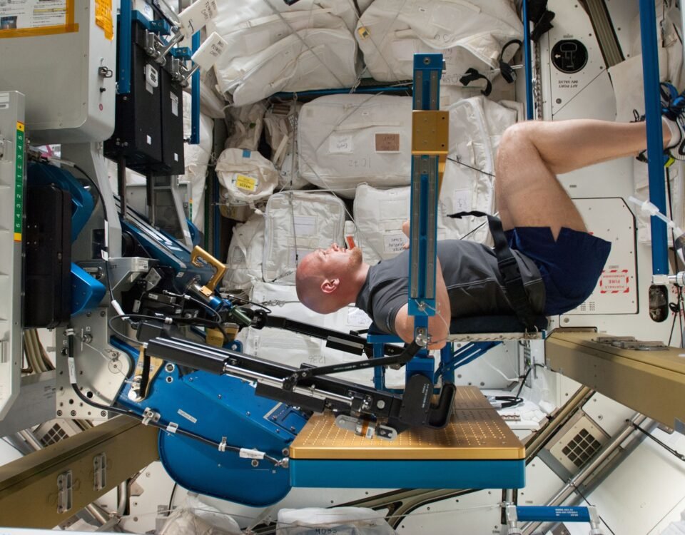 ESA astronaut Alexander Gerst in a squat position while working out on the ARED, with his arms against a beam. His body is facing to the right and his head turned to smile at the camera.