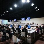 Leaders from two dozen space agencies sit in rows of chairs around a large open rectangular table illuminated under a truss of white lights.
