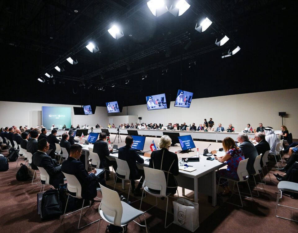 Leaders from two dozen space agencies sit in rows of chairs around a large open rectangular table illuminated under a truss of white lights.