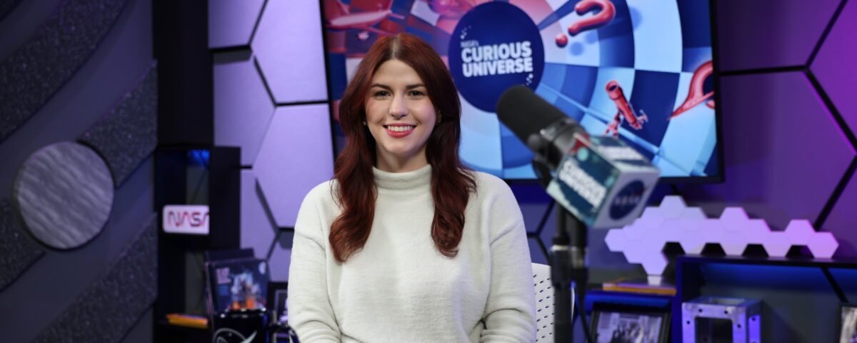 Katie Konans sits at a dark-colored desk wearing a white sweater with long brown hair. Behind her is a TV screen with the Curious Universe logo.