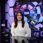Katie Konans sits at a dark-colored desk wearing a white sweater with long brown hair. Behind her is a TV screen with the Curious Universe logo.
